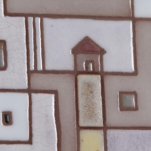 以下が含まれることがあります： 赤色の屋根と窓のある小さな家が描かれた幾何学模様のセラミックタイル。タイルは白、茶色、黄色など、さまざまな色の正方形と長方形で構成されています。
