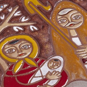 May include: A ceramic tile depicting a religious scene with figures in stylized form. The artwork features a woman holding a baby, a shepherd with a staff, and a bird in a tree, all rendered in earthy tones.