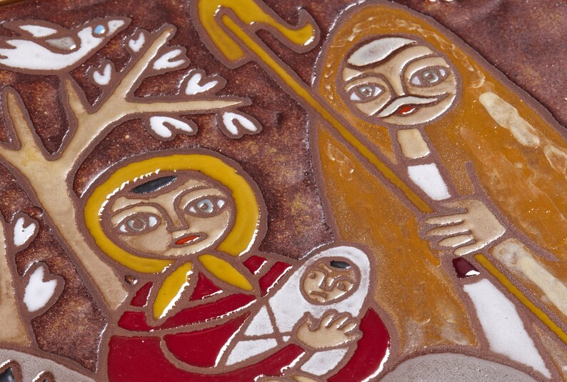 May include: A ceramic tile depicting a religious scene with figures in stylized form. The artwork features a woman holding a baby, a shepherd with a staff, and a bird in a tree, all rendered in earthy tones.