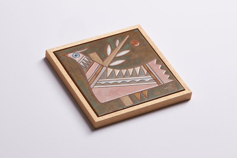 May include: A framed ceramic tile with a stylized pink bird with geometric patterns and a green branch with leaves on a brown background.