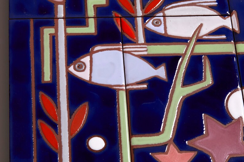 May include: A close-up of a blue and white ceramic tile with a stylized design of two fish swimming among red and green plants. The tile has a textured surface and a glossy finish.