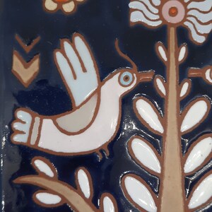 May include: A white bird with blue accents flies over a brown branch with white leaves against a dark blue background. The bird has a blue eye and a long beak.