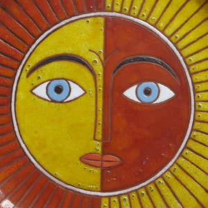 May include: A ceramic plate with a stylized sun face design. The sun has a yellow and orange face with blue eyes and a red mouth. The plate has a red and yellow rim.