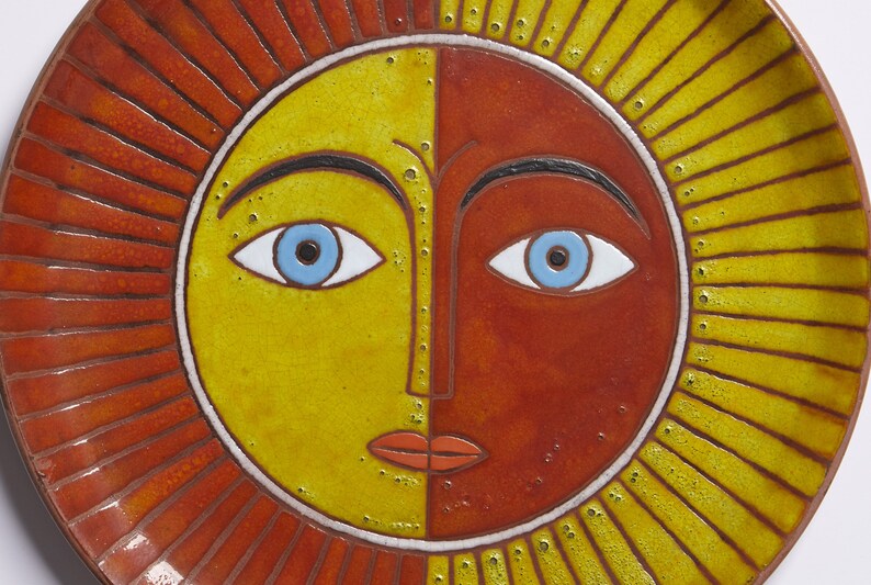 May include: A ceramic plate with a stylized sun face design. The sun has a yellow and orange face with blue eyes and a red mouth. The plate has a red and yellow rim.
