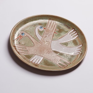 May include: A ceramic plate with a green glaze features a stylized design of two birds in flight. The birds are painted in white and brown with blue eyes.