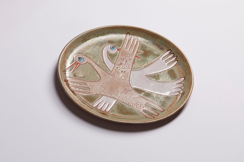 May include: A ceramic plate with a green glaze features a stylized design of two birds in flight. The birds are painted in white and brown with blue eyes.