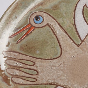 May include: A ceramic plate with a hand-painted design of two white birds with orange beaks and blue eyes. The plate has a green and brown glaze.