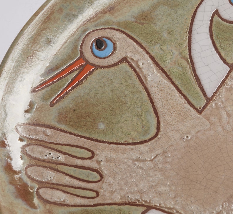 May include: A ceramic plate with a hand-painted design of two white birds with orange beaks and blue eyes. The plate has a green and brown glaze.
