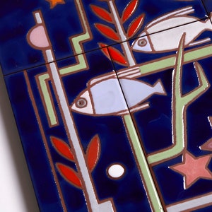 May include: A close-up of a blue ceramic tile with a geometric design featuring two fish, a star, and a plant. The fish are white and blue, the star is pink and red, and the plant is green and red.