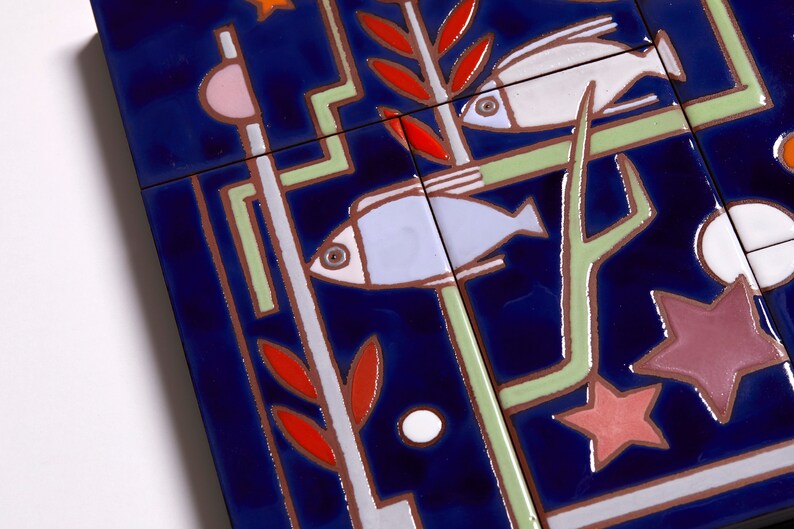 May include: A close-up of a blue ceramic tile with a geometric design featuring two fish, a star, and a plant. The fish are white and blue, the star is pink and red, and the plant is green and red.