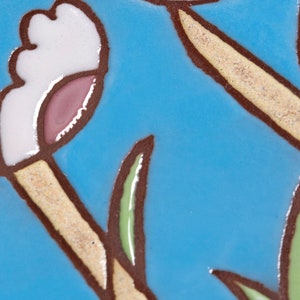 Puede incluir: Primer plano de un azulejo de cer&aacute;mica azul con un dise&ntilde;o de flor blanca y rosa. La flor tiene un tallo marr&oacute;n y hojas verdes.