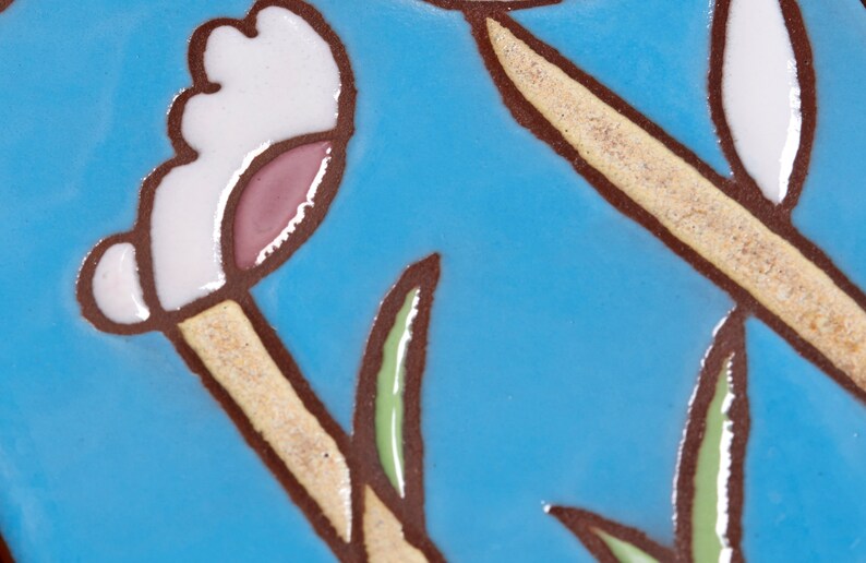 Puede incluir: Primer plano de un azulejo de cer&aacute;mica azul con un dise&ntilde;o de flor blanca y rosa. La flor tiene un tallo marr&oacute;n y hojas verdes.