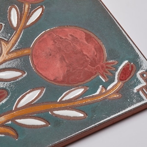 May include: A ceramic tile with a blue-green background featuring a stylized orange branch with white leaves and red pomegranates. The tile is signed "TIB" in white.
