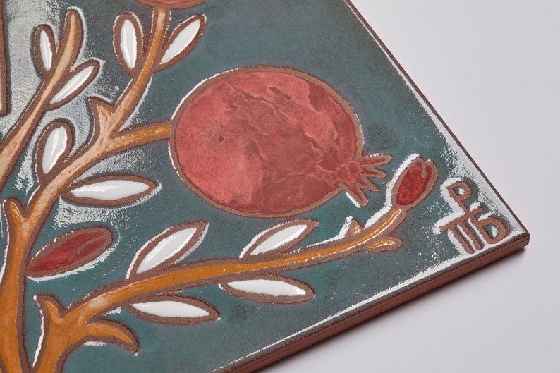 May include: A ceramic tile with a blue-green background featuring a stylized orange branch with white leaves and red pomegranates. The tile is signed "TIB" in white.