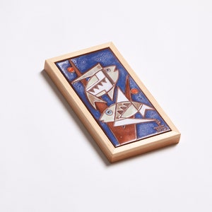 May include: A framed ceramic tile with a blue background and two stylized fish in white, brown, and orange. The fish are depicted swimming upwards, with their tails pointing towards the top of the tile. The tile is framed in a light wood frame.