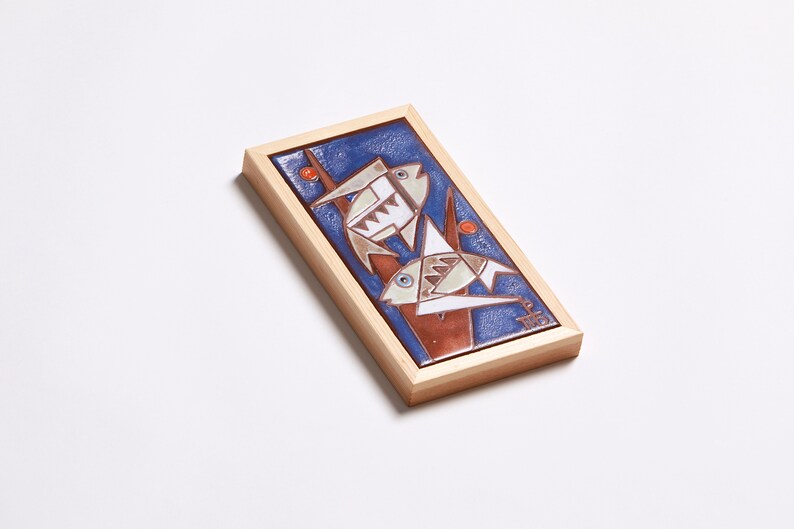 May include: A framed ceramic tile with a blue background and two stylized fish in white, brown, and orange. The fish are depicted swimming upwards, with their tails pointing towards the top of the tile. The tile is framed in a light wood frame.