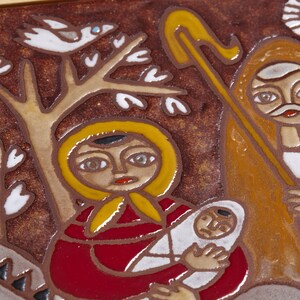 May include: A ceramic tile depicting a religious scene. A woman in a red robe and yellow head covering holds a baby wrapped in white. A figure with a staff and a sun are also present. The background is brown with a tree and a bird.