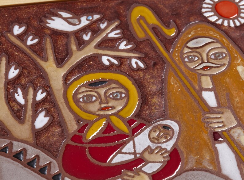 May include: A ceramic tile depicting a religious scene. A woman in a red robe and yellow head covering holds a baby wrapped in white. A figure with a staff and a sun are also present. The background is brown with a tree and a bird.