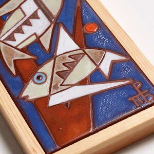 May include: A framed ceramic tile with a blue background and a stylized image of three fish in shades of brown, white, and blue. The tile is signed "MB" in the lower right corner.