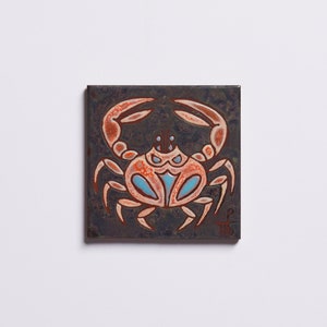 Puede incluir: Un azulejo de cerámica con un diseño de cangrejo naranja estilizado sobre un fondo gris oscuro. El cangrejo tiene detalles azules y está centrado en el azulejo.