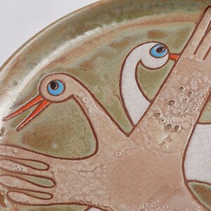May include: A ceramic plate with a hand-painted design of two white birds with blue eyes and orange beaks. The birds are flying with their wings outstretched. The plate has a green and brown glaze.