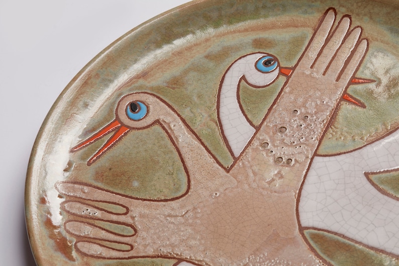 May include: A ceramic plate with a hand-painted design of two white birds with blue eyes and orange beaks. The birds are flying with their wings outstretched. The plate has a green and brown glaze.