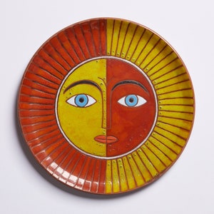 May include: A ceramic plate featuring a sun face design. The sun has a yellow and orange face with blue eyes and a red mouth. The plate has a red and yellow rim.