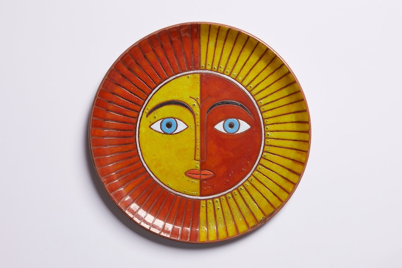 May include: A ceramic plate with a sun face design. The sun has a yellow and orange face with blue eyes and a red mouth. The plate has a red and yellow rim.