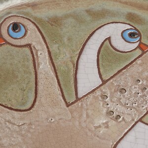 May include: A ceramic plate with a stylized design of two birds with outstretched wings. The birds are painted in shades of brown, white, and blue.