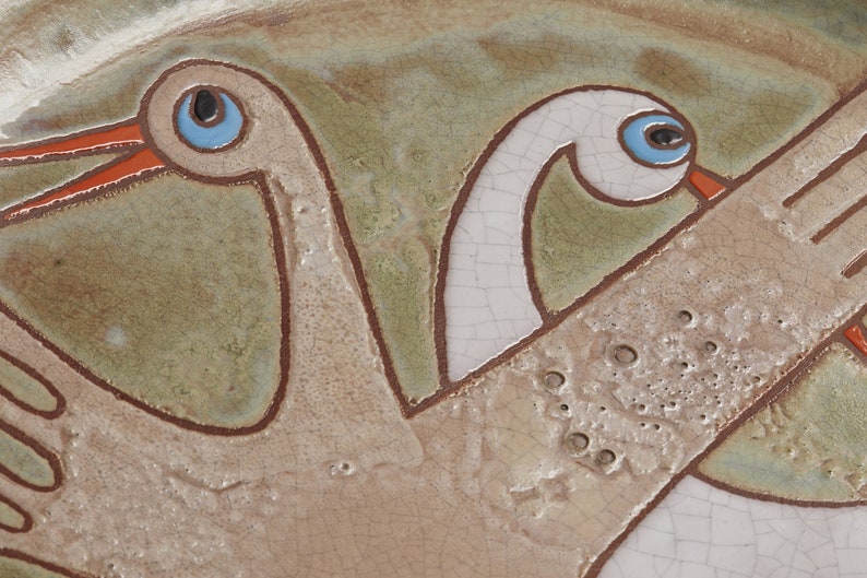 May include: A ceramic plate with a stylized design of two birds with outstretched wings. The birds are painted in shades of brown, white, and blue.