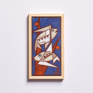 May include: A framed ceramic tile with a blue background and two white fish with geometric patterns. The fish are on a red and orange branch with orange dots. The tile is signed "P.T.B."