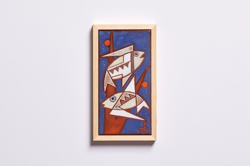 May include: A framed ceramic tile with a blue background and two white fish with geometric patterns. The fish are on a red and orange branch with orange dots. The tile is signed "P.T.B."