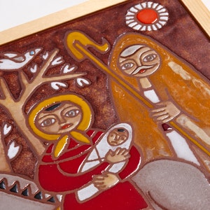 May include: A framed ceramic tile with a religious scene. The artwork features figures in red, yellow, and brown tones, with a baby held by a woman. A figure with a staff and a sun-like design are also present.