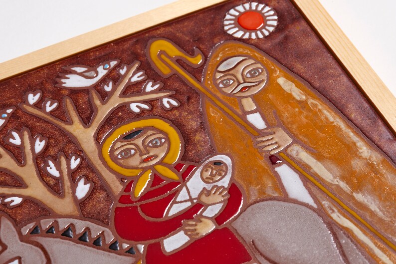 May include: A framed ceramic tile with a religious scene. The artwork features figures in red, yellow, and brown tones, with a baby held by a woman. A figure with a staff and a sun-like design are also present.