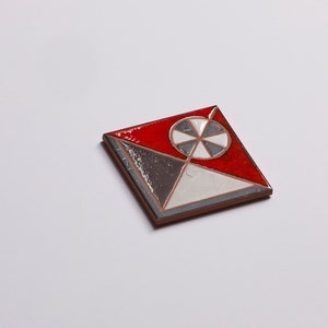 May include: A square ceramic tile with a geometric design. The tile is red, white, and gray with a black and white circle in the center.