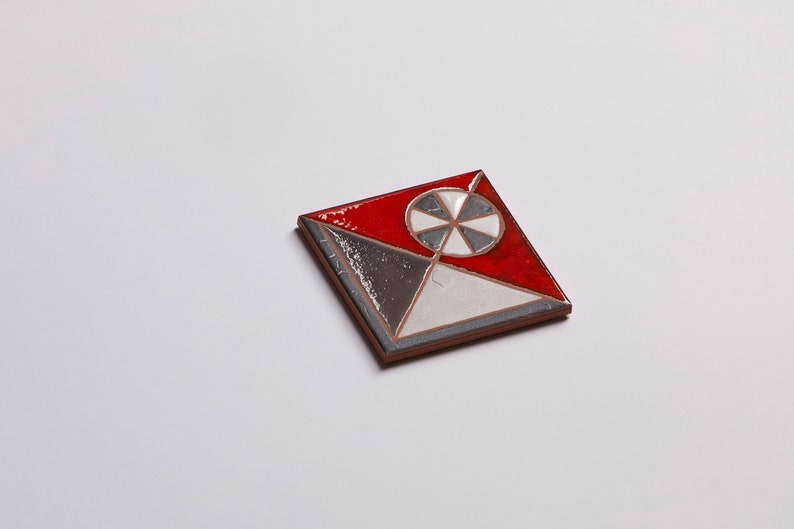 May include: A square ceramic tile with a geometric design. The tile is red, white, and gray with a black and white circle in the center.
