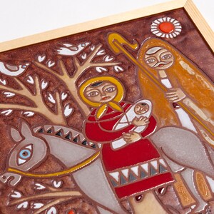 May include: A framed ceramic tile featuring a religious scene. The artwork depicts a woman in a red dress holding a baby, riding a donkey, with a man walking alongside. The background is brown with white birds and trees.