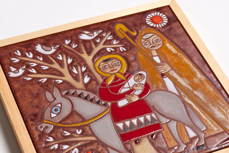 May include: A framed ceramic tile featuring a religious scene. The artwork depicts a woman in a red dress holding a baby, riding a donkey, with a man walking alongside. The background is brown with white birds and trees.