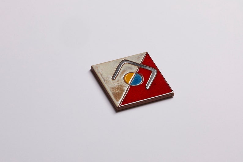 K&ouml;nnte beinhalten: Eine quadratische Keramikfliese mit einem rot-wei&szlig;-blauen Design. Die Fliese zeigt eine stilisierte geometrische Form mit einem Kreis in der Mitte.
