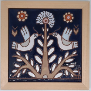 May include: A ceramic tile with a blue background features two white birds perched on a brown tree with white leaves. The tree has a white flower with a pink center. There are three white circles with orange and pink centers on the tile.