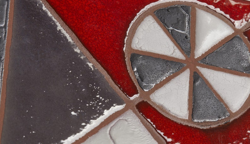 May include: Close-up of a red, white, and gray mosaic tile with a geometric design. The tile features a circular pattern with six spokes radiating from the center.