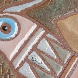 May include: A ceramic tile with a stylized bird design. The bird is depicted in shades of brown, pink, and blue, with a large blue eye. The tile has a textured surface and a rustic appearance.