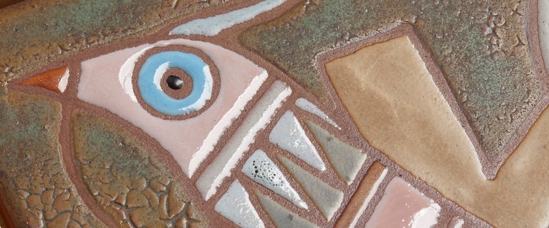 May include: A ceramic tile with a stylized bird design. The bird is depicted in shades of brown, pink, and blue, with a large blue eye. The tile has a textured surface and a rustic appearance.