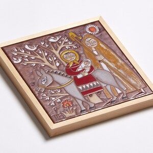 May include: A framed ceramic tile depicting a religious scene. The artwork features figures on a donkey, with a tree, birds, and a sun. The color palette includes brown, red, yellow, and white, set within a light wood frame.