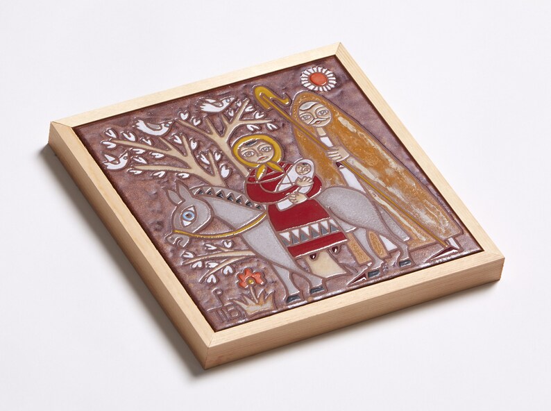 May include: A framed ceramic tile depicting a religious scene. The artwork features figures on a donkey, with a tree, birds, and a sun. The color palette includes brown, red, yellow, and white, set within a light wood frame.