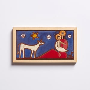 May include: A framed ceramic tile with a blue background depicting a white cow, a red hill, a yellow star, and three figures in white and yellow. The figures are standing on the hill and appear to be a family.