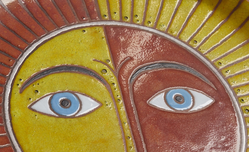 May include: A ceramic sun face with a yellow and red background. The sun has two blue eyes with white pupils and black outlines. The sun has a black eyebrow above each eye.