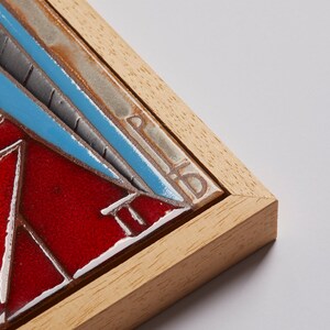 May include: A framed ceramic tile with a geometric design in shades of red, blue, and gray. The tile is framed in a light wood frame.