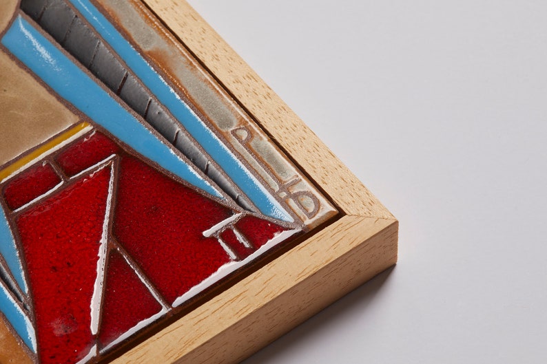 May include: A framed ceramic tile with a geometric design in shades of red, blue, and gray. The tile is framed in a light wood frame.