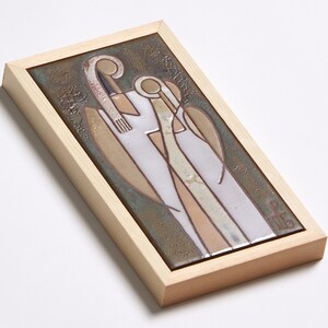 May include: A framed ceramic tile with a stylized image of two figures in white, brown, and beige against a green background. The figures are standing close together and appear to be embracing. The tile is framed in a light wood frame.